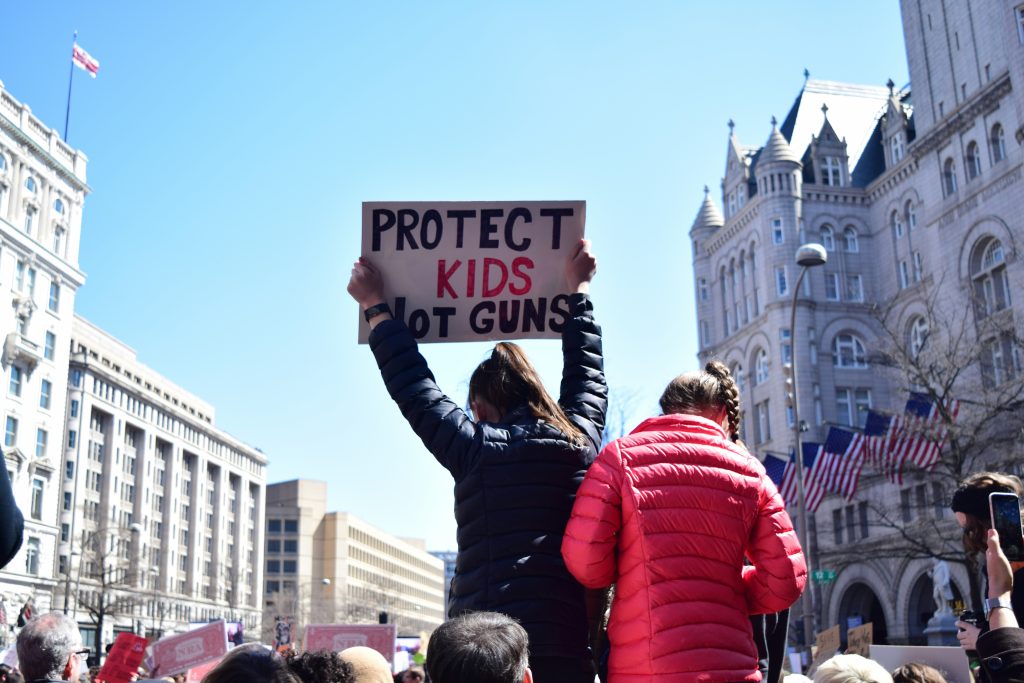 A protest with a person holding a sign saying "Protect kids not guns"